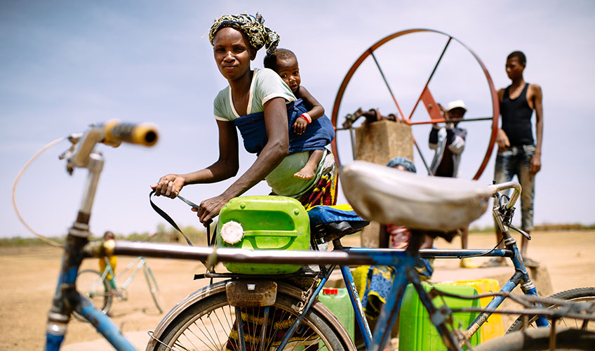 Woman with baby on her back untying a jerry can from a bicycle as she prepares to collect water. Two other people stand behind her at a pump.