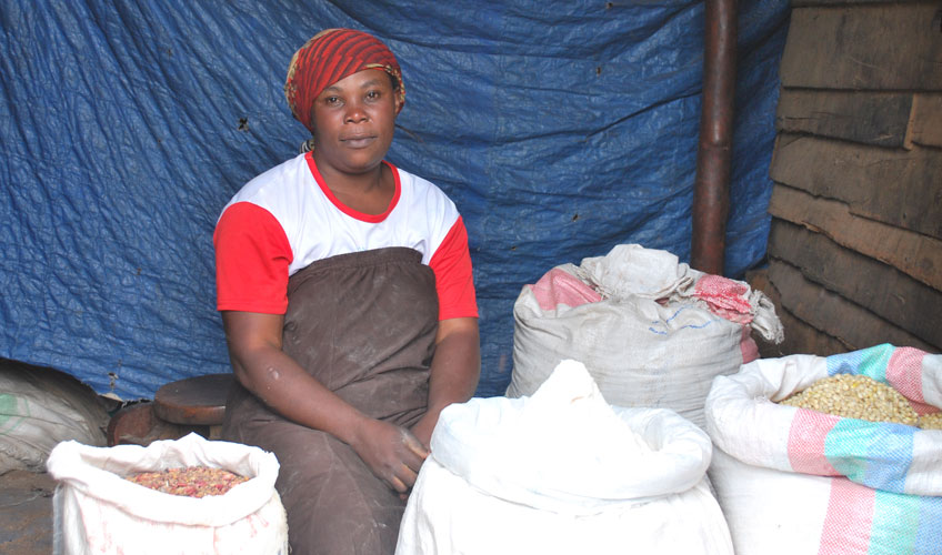 Safari wears a red hat and white t-shirt with red short sleeves. She is sitting on a small wooden stall surrounded by large white sacks of corn and flour. A blue plastic tarpaulin sheet hangs in the background.