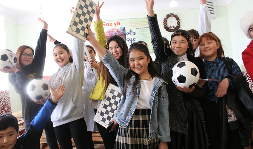 A group of children celebrate the opening of the children's development centre. They raise their hands in the air holding footballs and games and smiling. 