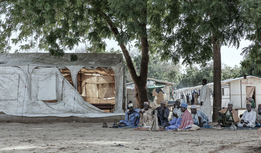 Group of men seated under trees in a displacement camp.