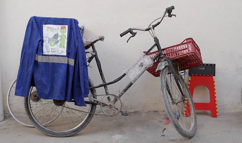 Bicycle propped up against the wall, with a rubbish collector's jacked hanging from the seat and a plastic basket over the front wheel.