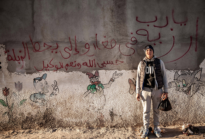 A young man stands in front of a graffitied wall with a plastic bag in hand