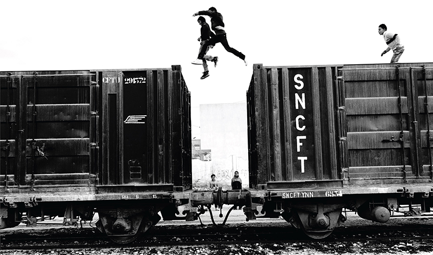 Black and white photo showing young people running along the top of a freight train with two of them caught mid-air jumping between containers.