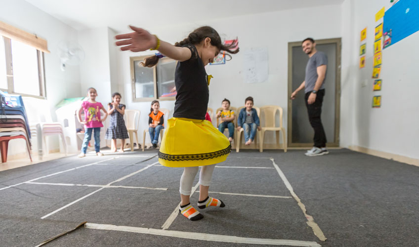 A girl in a bright yellow skirt jumps through squares marked on the floor as other children and an adult look on.