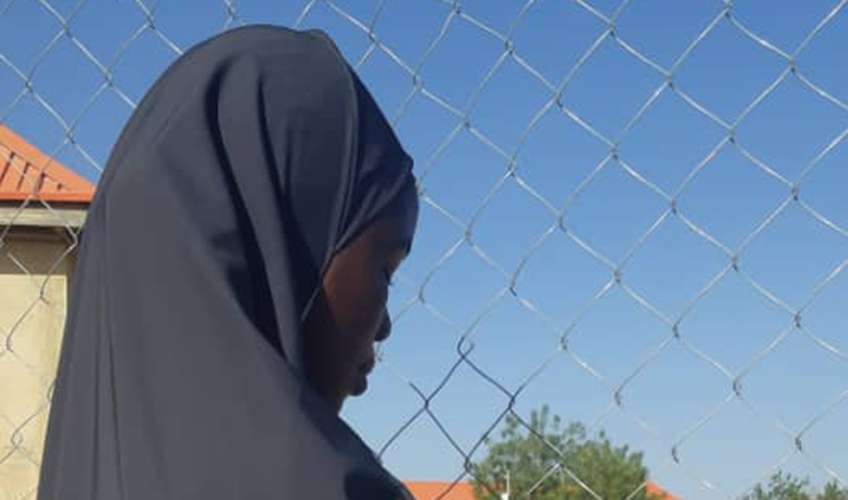 A girl with a headscarf and her back to the camera looks sadly at a wire fence. Blue skies and trees can be seen in the background.
